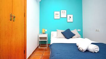A room with a wooden door, a bed with a blue blanket and white pillows, a white bedside table with a lamp, and a wall with three framed pictures.