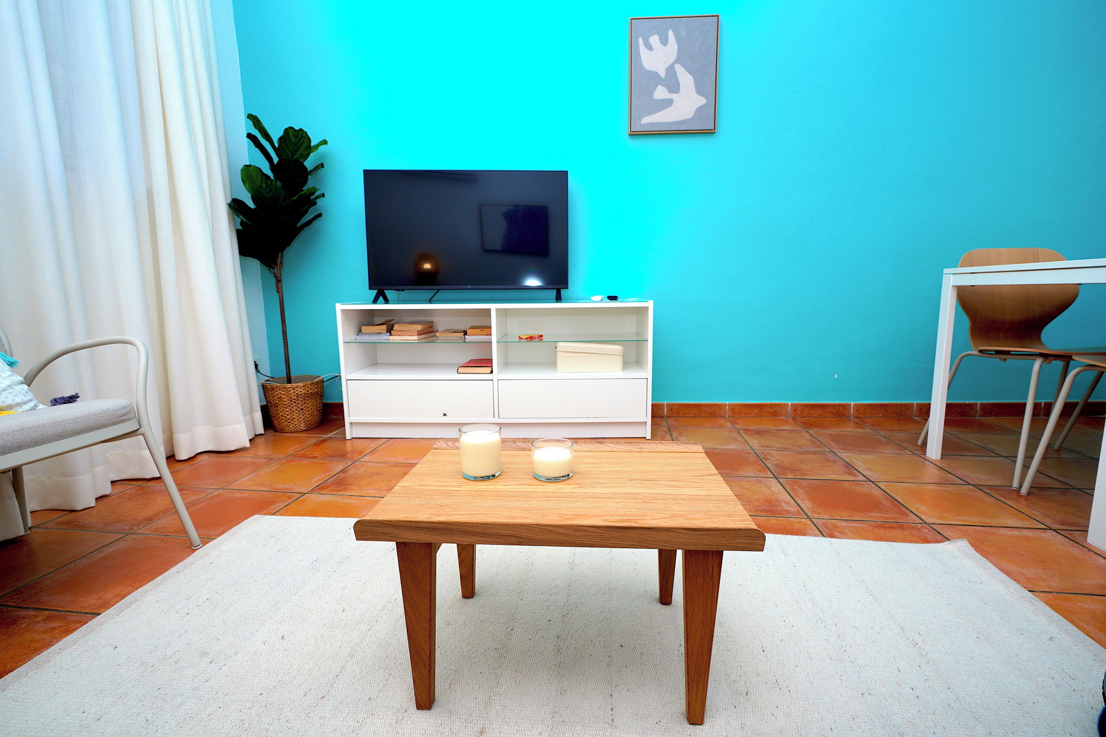 Una sala de estar con televisor, un mueble blanco, una mesa de madera y una planta.