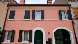 Un edificio de dos pisos con fachada color melocotón, contraventanas verdes y una puerta central arqueada.