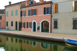 A canal-side building with arched doorways, a bicycle, and a blue boat. - GuestHost - A comfortable and welcoming 75 square meters apartment with a large courtyard and private entrance, perfect for six people, located on the ground floor of a renovated historic building facing the canal (NOT accessible by car).The property enj (Comacchio)