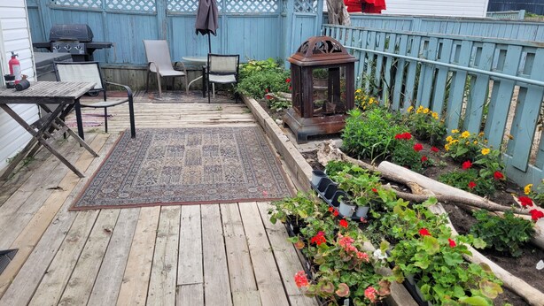 A wooden deck with a grill, chairs, and a table. There is a garden with flowers and a bird feeder.