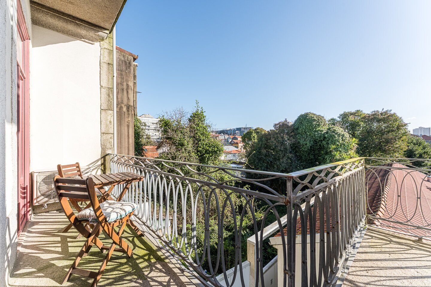 Un balcon avec une table et des chaises, une rambarde en métal et une vue sur des immeubles et des arbres.