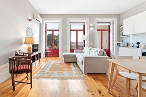 A modern apartment with a living room, dining area, and kitchen. It features a large window, a red door, and a wooden floor. - GuestReady - Historic charm with a balcony (Porto)