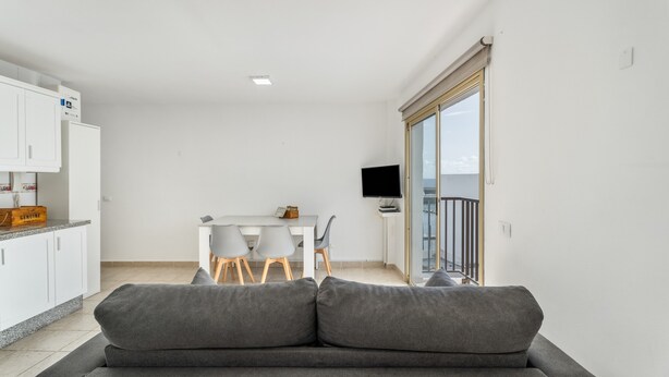 Una sala de estar moderna con un sofá gris, un comedor con mesa y sillas, una cocina con armarios blancos y un televisor sobre un mueble.