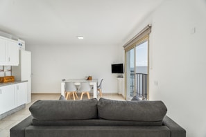 Un salon moderne avec un canapé gris, un coin repas avec une table et des chaises, une cuisine avec des meubles blancs et un téléviseur posé sur un meuble.