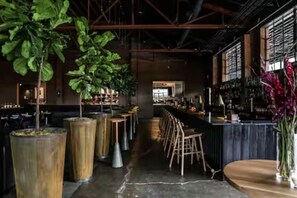 Uma área de bar com pé direito alto, vigas aparentes e grandes vasos de plantas.