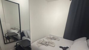 Ein kleines Hotelzimmer mit einem Einzelbett, einem kleinen Schreibtisch, einem Stuhl, einem Spiegel und einem Fenster mit Vorhang.