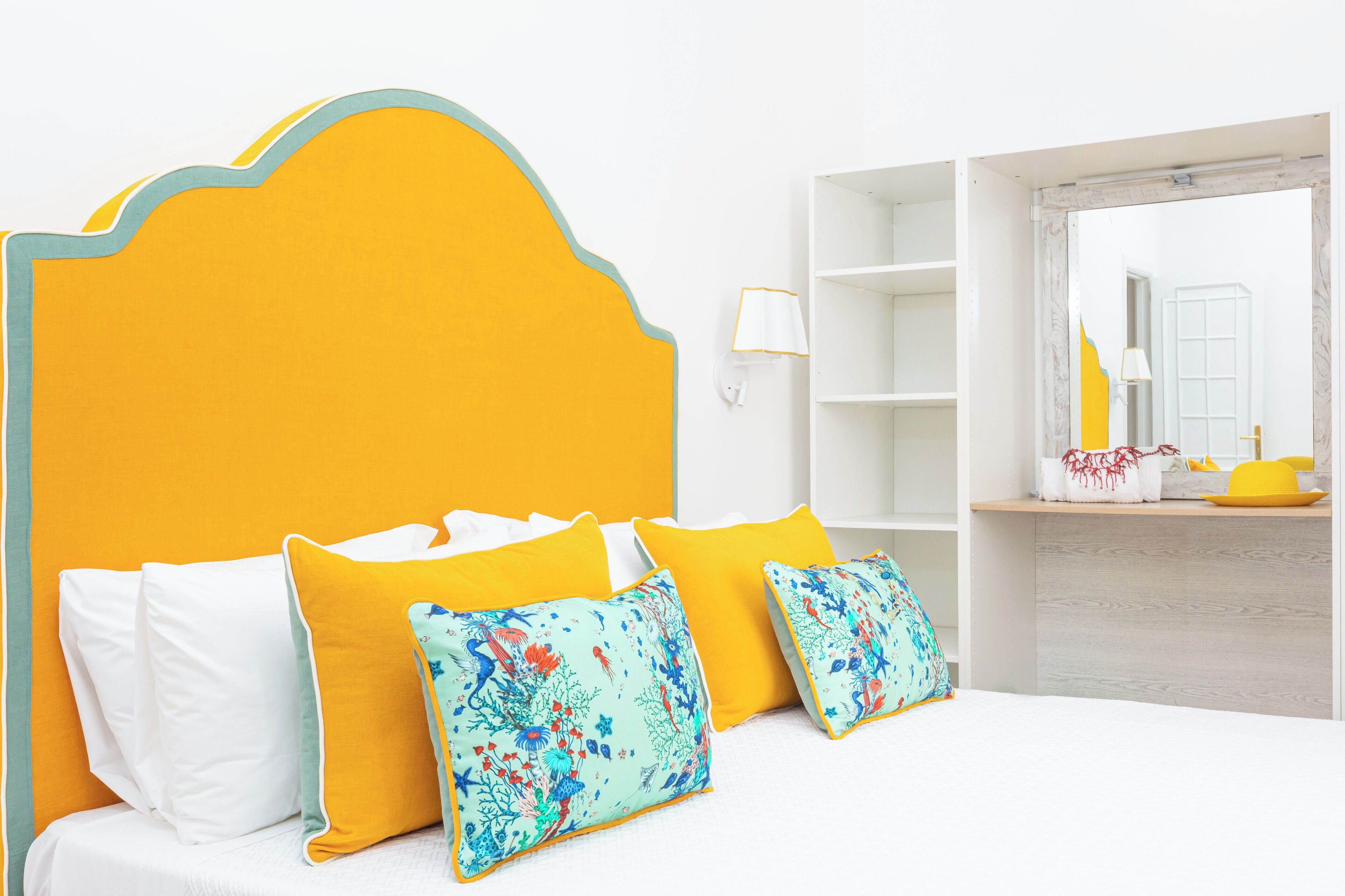 Un lit avec une tête de lit jaune, des oreillers blancs et jaunes, et un coussin à motifs floraux. Il y a un mur blanc avec une étagère et une lampe.