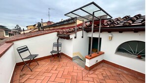 Une terrasse sur le toit, dotée d’un sol carrelé, de deux chaises, d’une petite table, d’une structure vitrée et offrant une vue sur les bâtiments voisins.