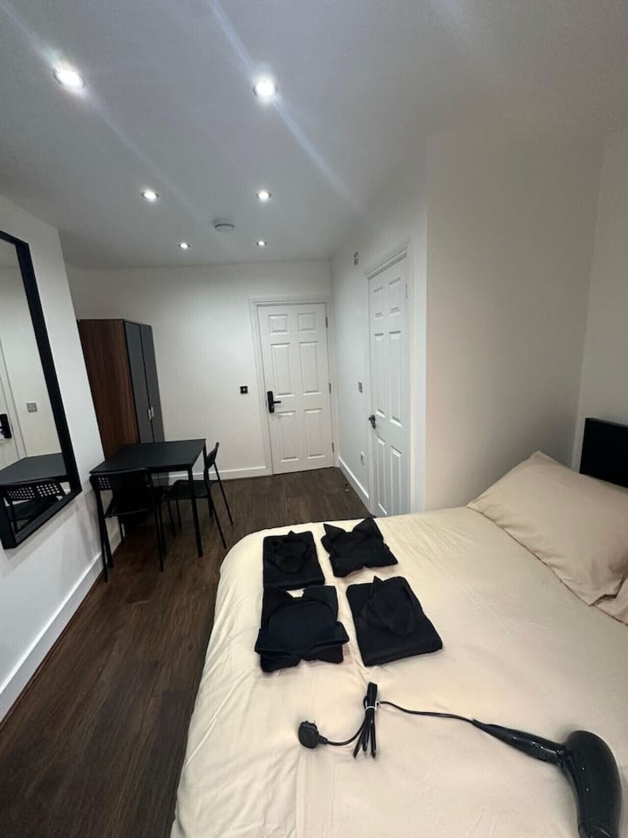 A bedroom with a bed, a desk, and a chair. There are clothes on the bed and a hairdryer on the floor.