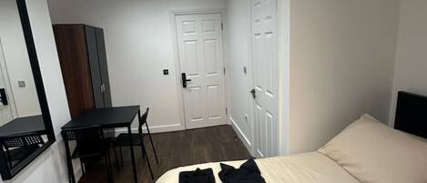 A bedroom with a bed, a desk, and a chair. There are clothes on the bed and a hairdryer on the floor.