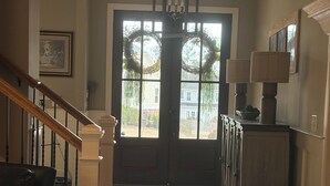 Un pasillo con suelo de madera, una escalera con barandilla blanca, un candelabro y una puerta decorada con coronas.