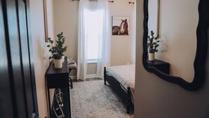 A bedroom with a bed, a bedside table with a plant, a window with curtains, and a wall-mounted mirror.