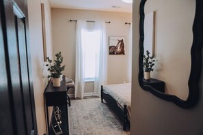 Un dormitorio con cama, mesita de noche con planta, ventana con cortinas y espejo adosado a la pared.