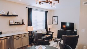 A modern living room with a dining table, chairs, a sofa, a TV, and a kitchen area with cabinets and a countertop.