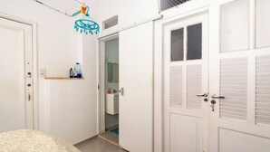 Ein Schlafzimmer mit einem Bett, einer weißen Tür, einem Regal mit Pflegeprodukten und einem durch eine offene Tür einsehbaren Badezimmer.