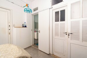 Ein Schlafzimmer mit einem Bett, einer weißen Tür, einem Regal mit Pflegeprodukten und einem durch eine offene Tür einsehbaren Badezimmer.