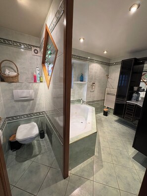 Ein Badezimmer mit Badewanne, Toilette und Waschbecken. Auf dem Waschbecken stehen Pflegeprodukte und an der Wand ein Korb.