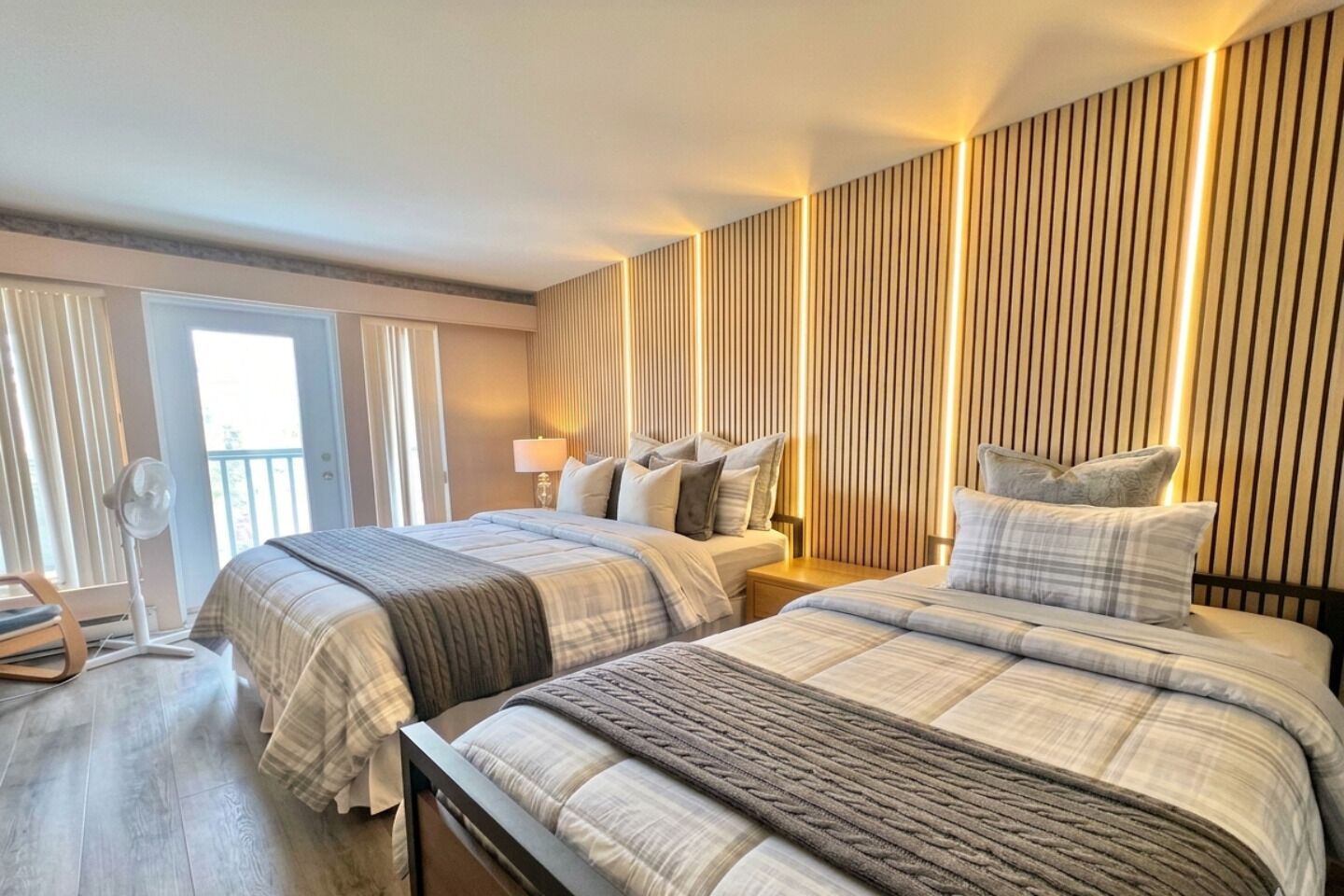 Habitación de hotel con dos camas, un balcón, un ventilador y una pared con líneas verticales.