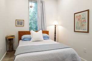 Un dormitorio con una cama, una lámpara de noche, una ventana con cortinas y un cuadro en la pared.