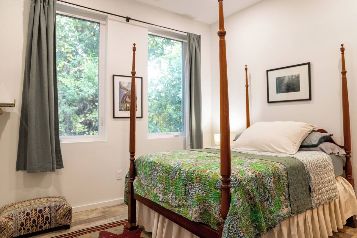 Una cama con dosel con un edredón estampado verde, un banco de madera y una ventana con cortinas.