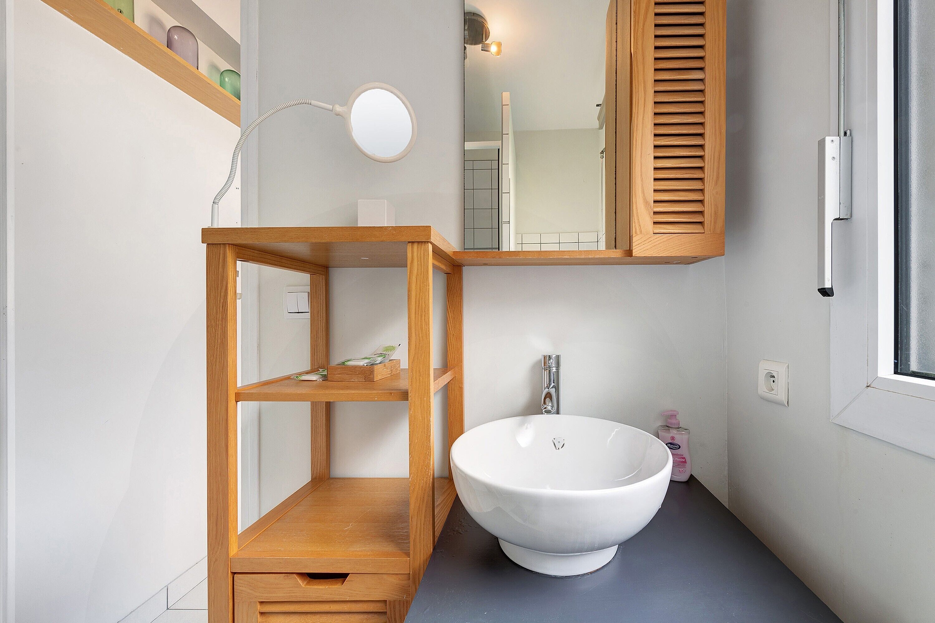 Un baño con un lavamanos blanco, estantes de madera, un espejo y una ventana.