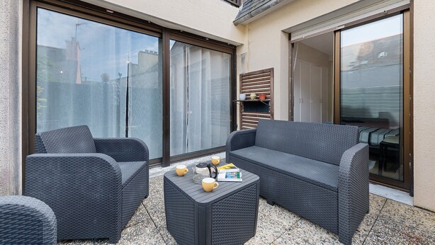 Un patio con sofá, sillón y mesa de centro, con puerta corredera de vidrio y vista a la calle.