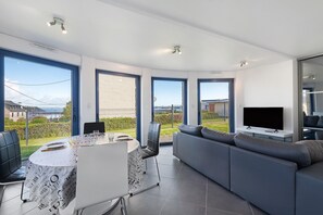 A modern living room with a dining area, large windows, and a flat-screen TV. - Serenity at Land’s End: Ocean Views and Beaches (Camaret-sur-Mer)