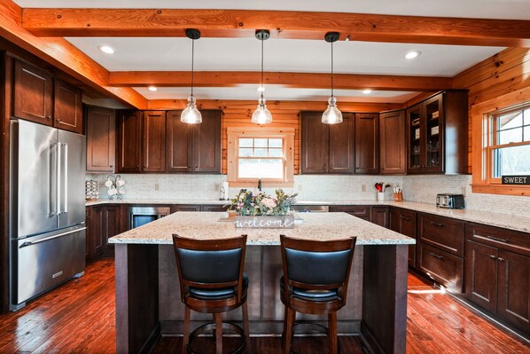 A kitchen with wooden cabinets, a stainless steel refrigerator, a marble island with two bar stools, pendant lights, and a window.