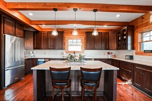 A kitchen with wooden cabinets, a stainless steel refrigerator, a marble island with two bar stools, pendant lights, and a window.