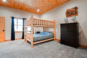 A bunk bed with a blue mattress, a wooden dresser, a window with blue curtains, and a wooden ceiling.
