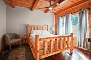 Ein hölzernes Bett mit Kopfteil, ein grau gepolsterter Sessel, ein kleiner Nachttisch mit einer Vase und ein Fenster mit Vorhängen.