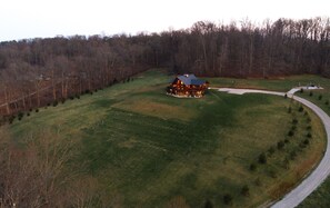 A large, single-story house with a wrap-around porch, surrounded by a grassy field and a forested area.