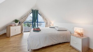 Un dormitorio con techo inclinado, una cama con colcha a rayas, una mesita de noche con lámpara y vistas a través de una ventana con cortinas.