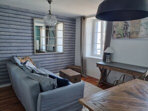 Une pièce avec un parquet en bois disposé en motif «chevrons», un canapé gris agrémenté de coussins bleus, une table en bois avec une lampe, une carte accrochée au mur et une fenêtre avec des rideaux.