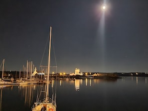Un port tranquille, de nuit, avec un voilier bien visible au premier plan, un bâtiment illuminé à l’arrière-plan et une pleine lune dans le ciel.