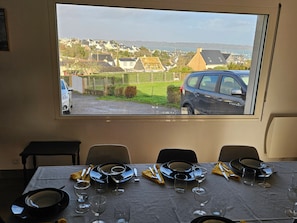 A dining table set with plates, glasses, and cutlery, with a view of houses and a car through a window. - House Overlooking Camaret with Harbor View (Camaret-sur-Mer)