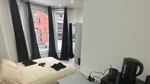 Et kompakt hotelværelse med seng, et lille tekøkken, et vindue med gardiner og udsigt til gaden.