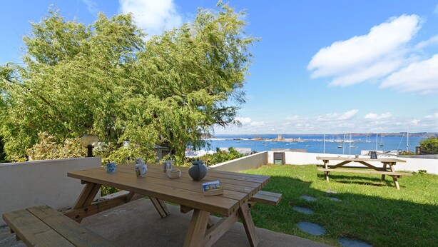 Une table de pique-nique en bois avec des bancs, un livre, une tasse et un vase, le tout placé sur une terrasse donnant sur un plan d’eau où naviguent des bateaux.