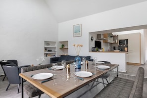 Un comedor moderno con una mesa larga dispuesta para cuatro personas, una cocina al fondo y un sofá gris.