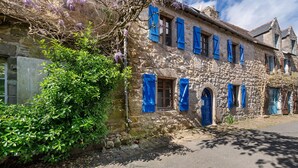 Um edifício de pedra com portadas azuis, uma rua calçada e uma árvore com flores roxas.