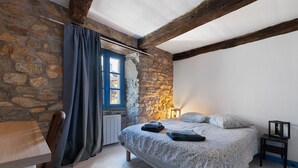 Quarto com parede de pedra, cama com roupa de cama estampada, janela com cortinas, uma pequena mesa e uma cadeira.