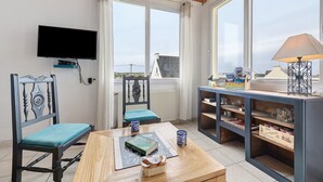 A room with a TV mounted on the wall, a table with a book and a mug, two chairs with blue cushions, and a window with a view of buildings. - Sea View, Authentic Fisherman's House (Camaret-sur-Mer)