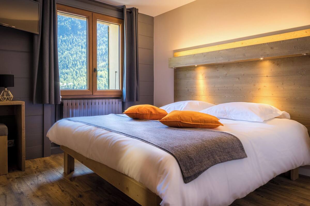 A neatly made bed with white and orange pillows, a wooden headboard, and a window with curtains.