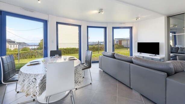 A modern living room with a large TV, grey sofa, and a dining area with a table and chairs. - Magical Moments by the Sea (Camaret-sur-Mer)