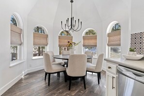 Un comedor con una mesa redonda y seis sillas, ventanas grandes con persianas, una araña y una zona de cocina con lavavajillas.