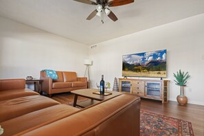 Una sala de estar con un sofá de cuero marrón, una mesa de centro, un televisor sobre un mueble de madera y un ventilador de techo.