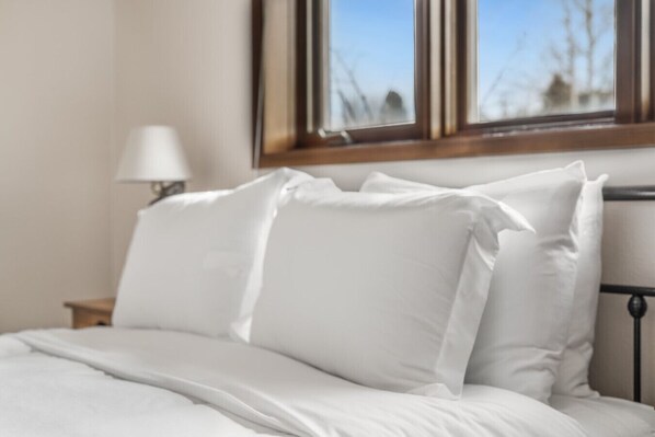 Una cama bien hecha con ropa blanca y almohadas, una lámpara de noche y una ventana con vista a árboles.