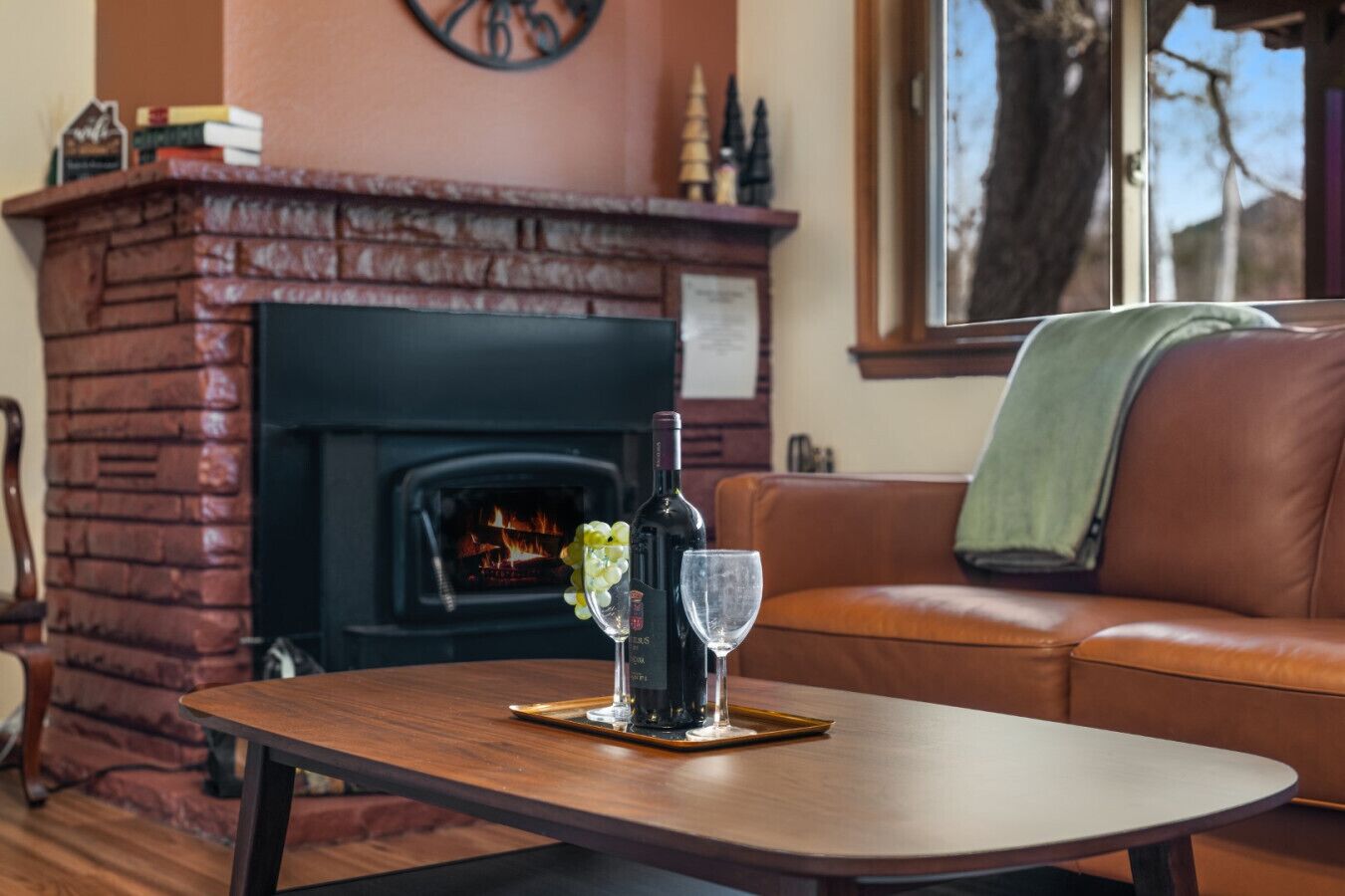 A cozy living room with a brick fireplace, a leather sofa, and a wooden coffee table with a bottle of wine and a glass.