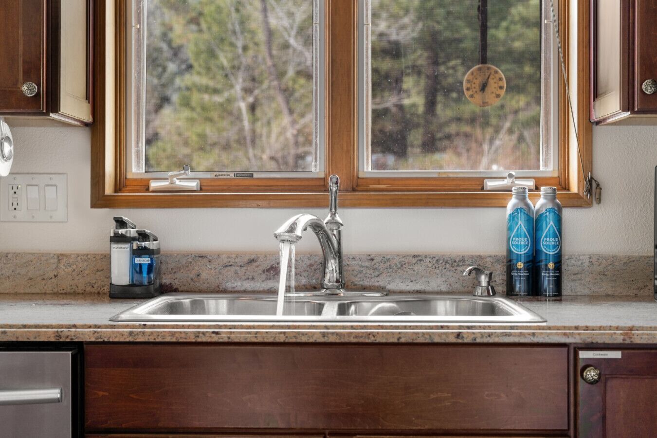 A kitchen with a double sink, wooden cabinets, a window with a view of trees, and two bottles of water on the counter.